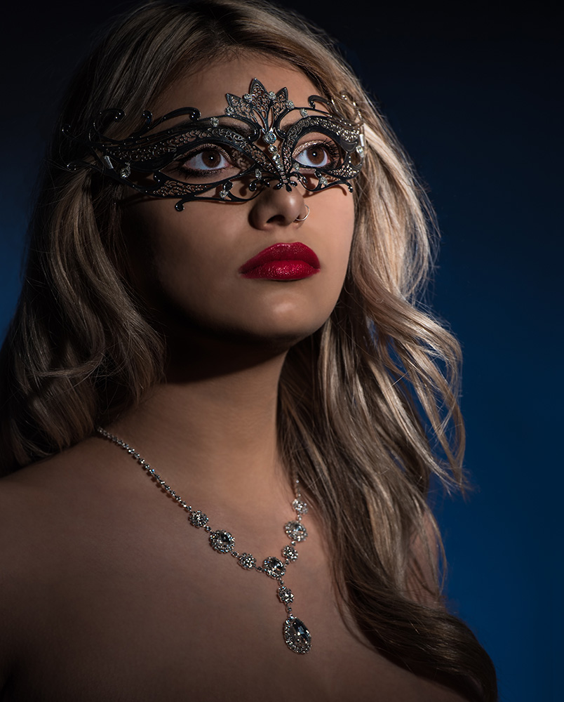 Blonde hair brown eye girl wearing red lipstick and a silver mascarade mask with a diamond neckless on a blue background. Alyssa Bryn Photography. Bryssa. Bryssa Blog.