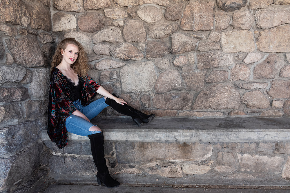 Blonde curly haired girl sits on bench by natural rock wall wearing knee high black boots, ripped skinny jeans, black lace bodysuite, and red flower patterned jacket. Alyssa Bryn Photography. Bryssa. Bryssa Blog.