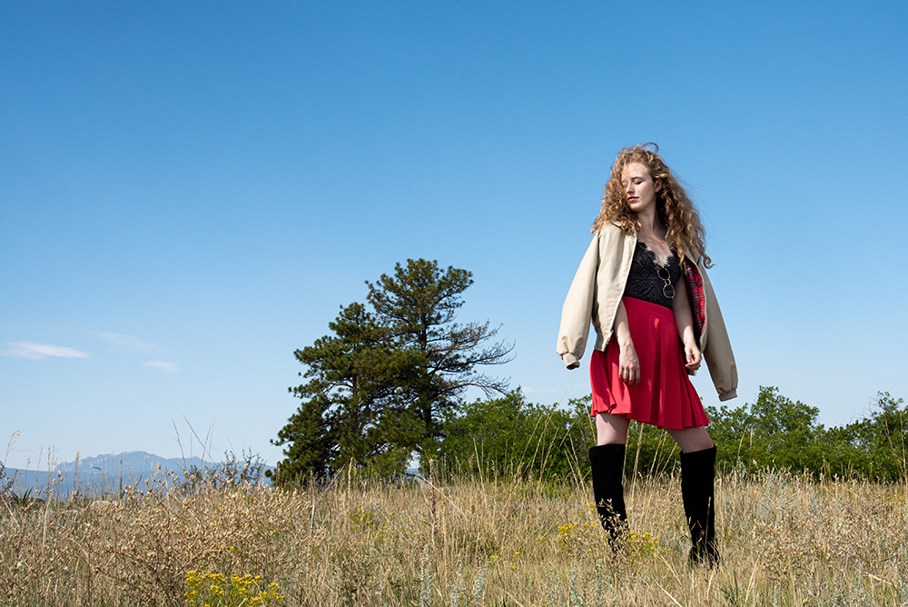 Girl standing in grass field with trees in the background wearing knee high black boots, pink skirt, black lace bodysuit and beige jacket over the shoulders. Alyssa Bryn Photography. Bryssa. Bryssa Blog.
