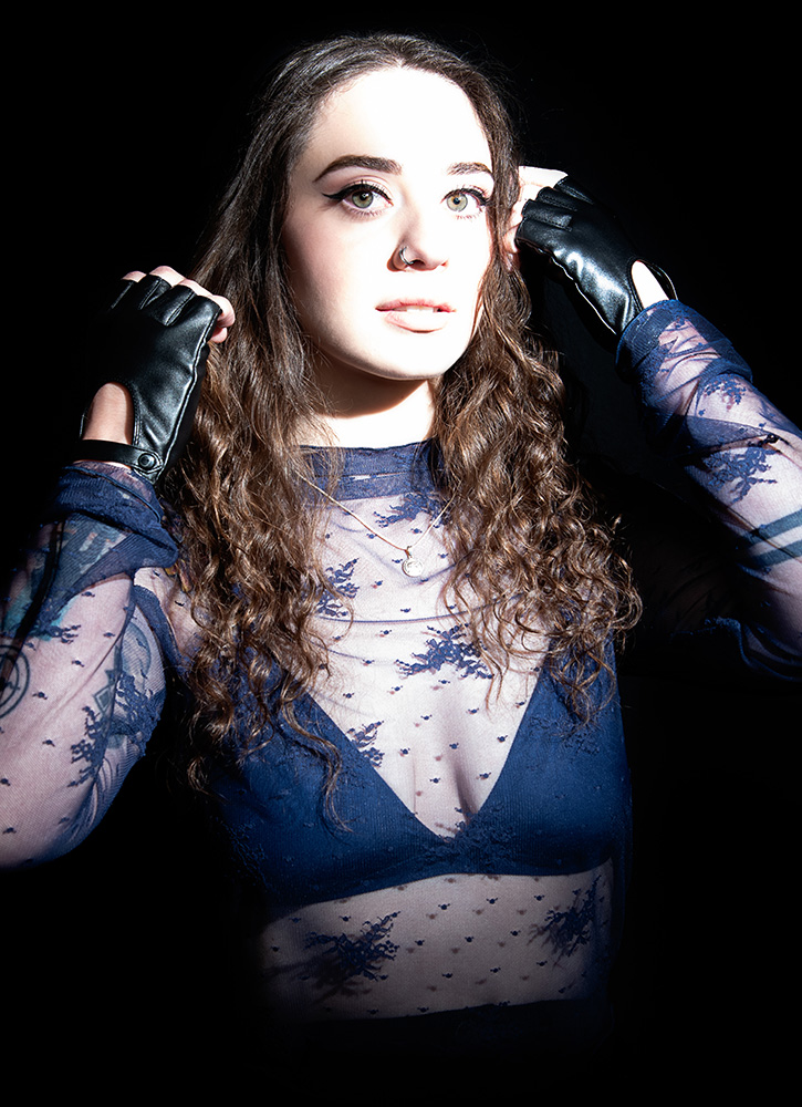 Tattooed girl with long drak curl hair wearing sheer midnight blue top with matching dark blue bra under, accessorizing with black leather gloves, silver neckless and nose ring, in a bright white light. Alyssa Bryn Photography. Bryssa. Bryssa Blog.