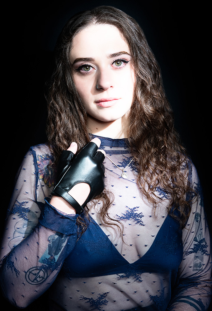 Tattooed girl with long drak curl hair wearing sheer midnight blue top with matching dark blue bra under, accessorizing with black leather gloves, silver neckless and nose ring, in a bright white light. Alyssa Bryn Photography. Bryssa. Bryssa Blog.