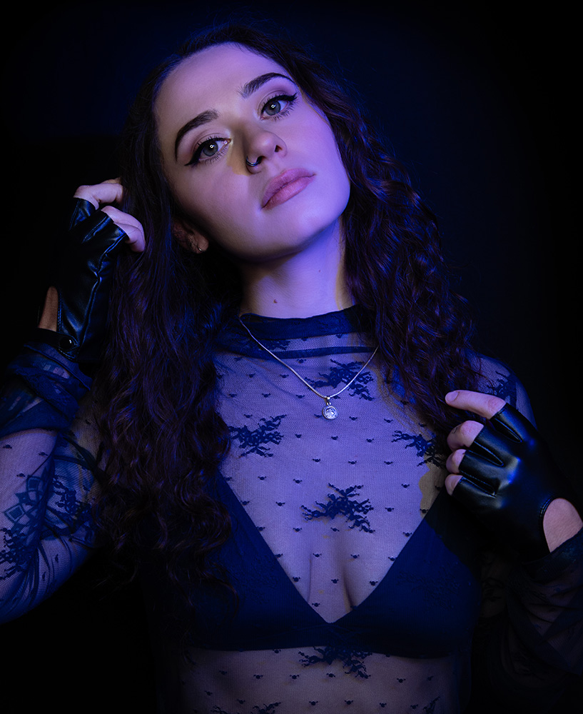 Tattooed girl with long drak curl hair wearing sheer midnight blue top with matching dark blue bra under, accessorizing with black leather gloves, silver neckless and nose ring, in a bright blue light. Alyssa Bryn Photography. Bryssa. Bryssa Blog.