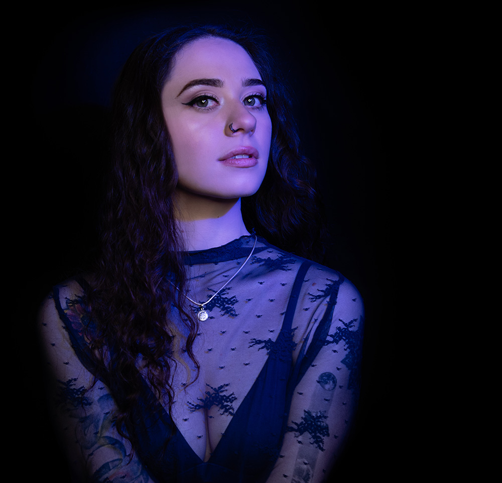  Tattooed girl with long drak curl hair wearing sheer midnight blue top with matching dark blue bra under, accessorizing with silver neckless and nose ring, in a bright blue light. Alyssa Bryn Photography. Bryssa. Bryssa Blog.