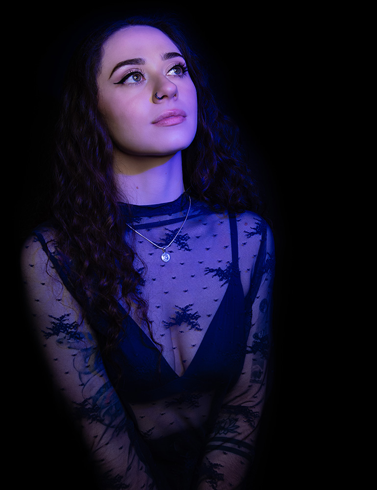 Tattooed girl with long drak curl hair wearing sheer midnight blue top with matching dark blue bra under, accessorizing with silver neckless and nose ring, in a bright blue light. Alyssa Bryn Photography. Bryssa. Bryssa Blog.